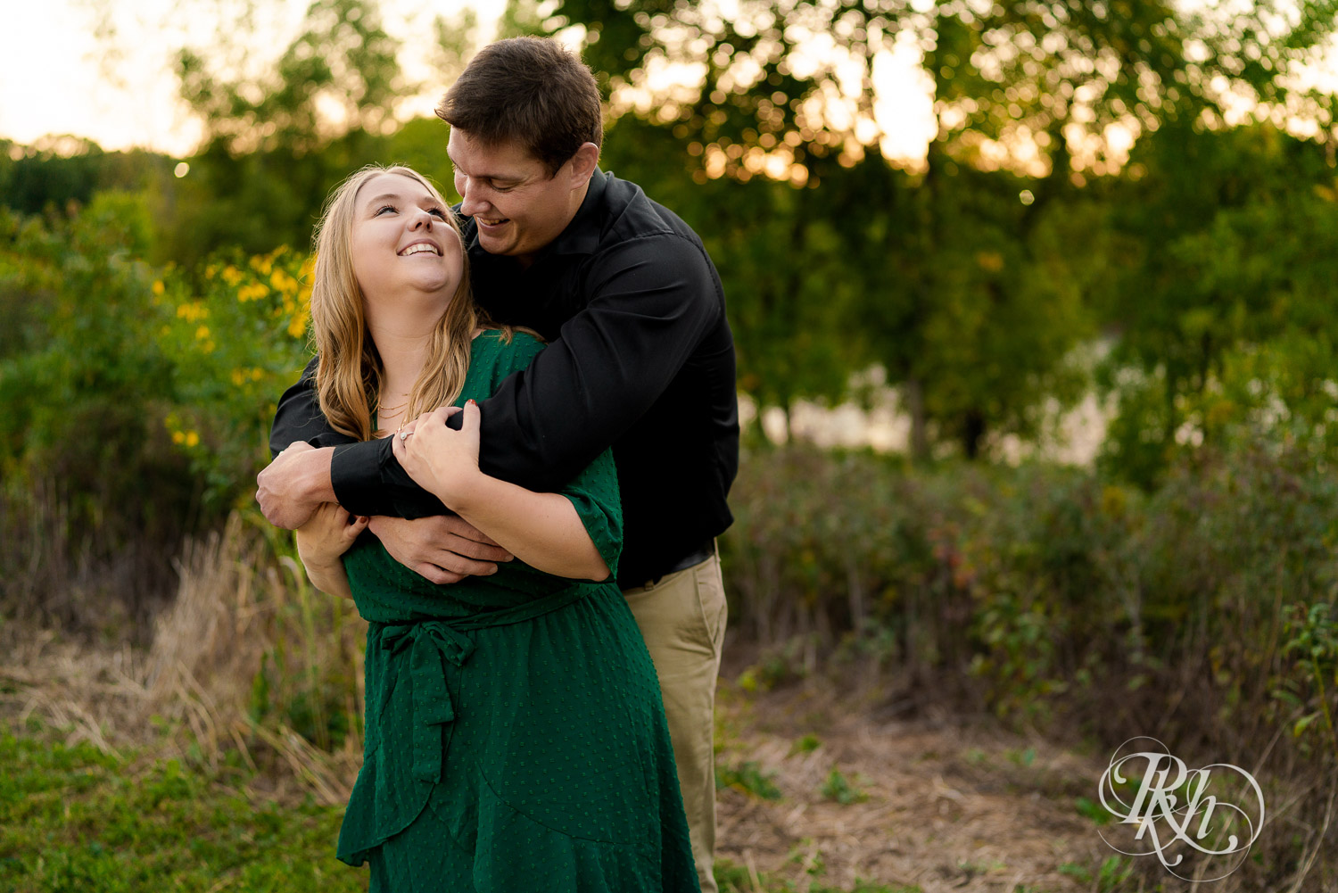 Man in black dress shirt hugging woman in green dress in front of trees and flowers in Eagan, Minnesota.