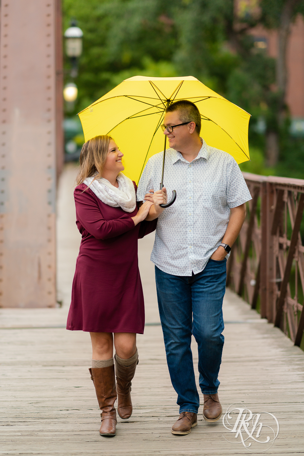 Man in glasses and woman is red dress and scarf smiling and holding umbrella on bridge in Minneapolis, Minnesota.