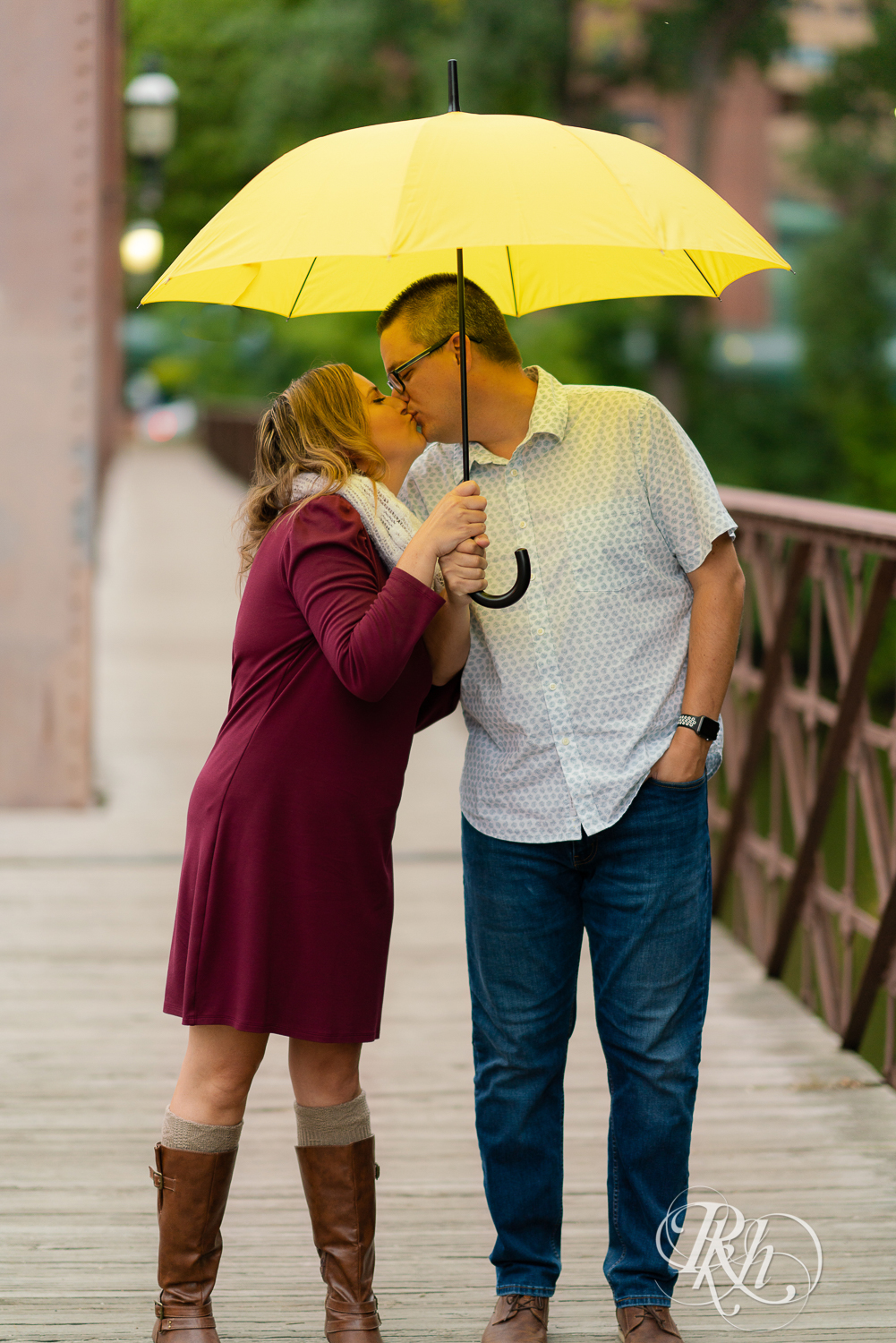 Man in glasses and woman is red dress and scarf kissing and holding umbrella on bridge in Minneapolis, Minnesota.