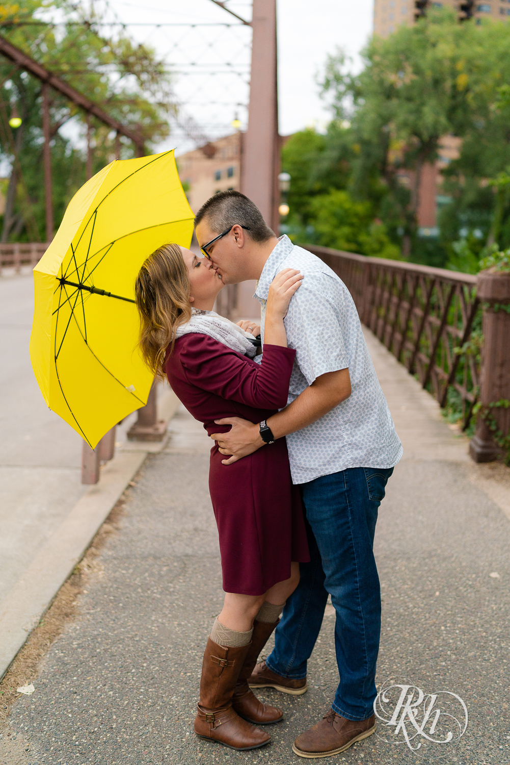 Man in glasses and woman is red dress and scarf kissing and holding umbrella on bridge in Minneapolis, Minnesota.