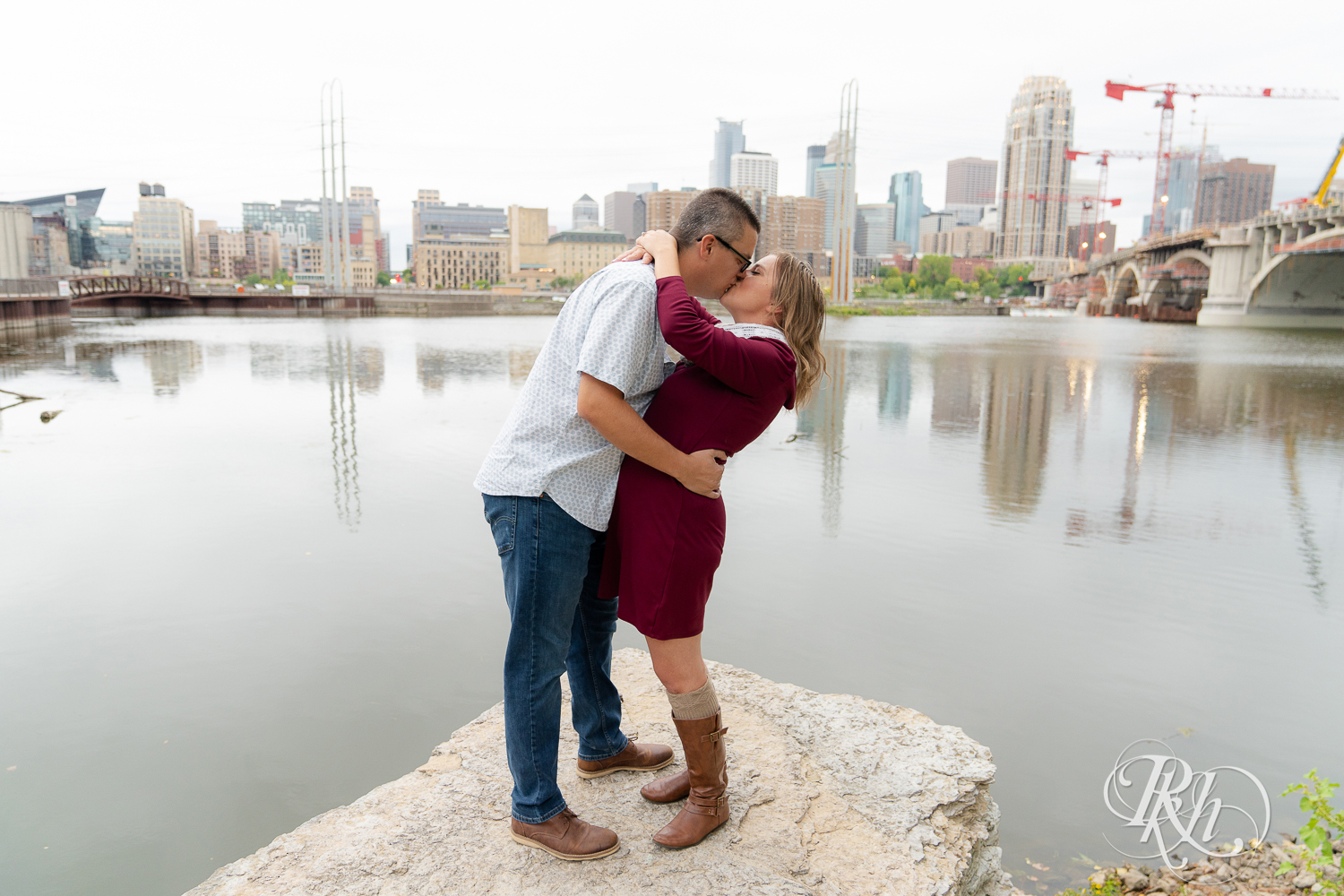 Man in glasses and woman is red dress and scarf kissing in Minneapolis, Minnesota with skyline in the background.