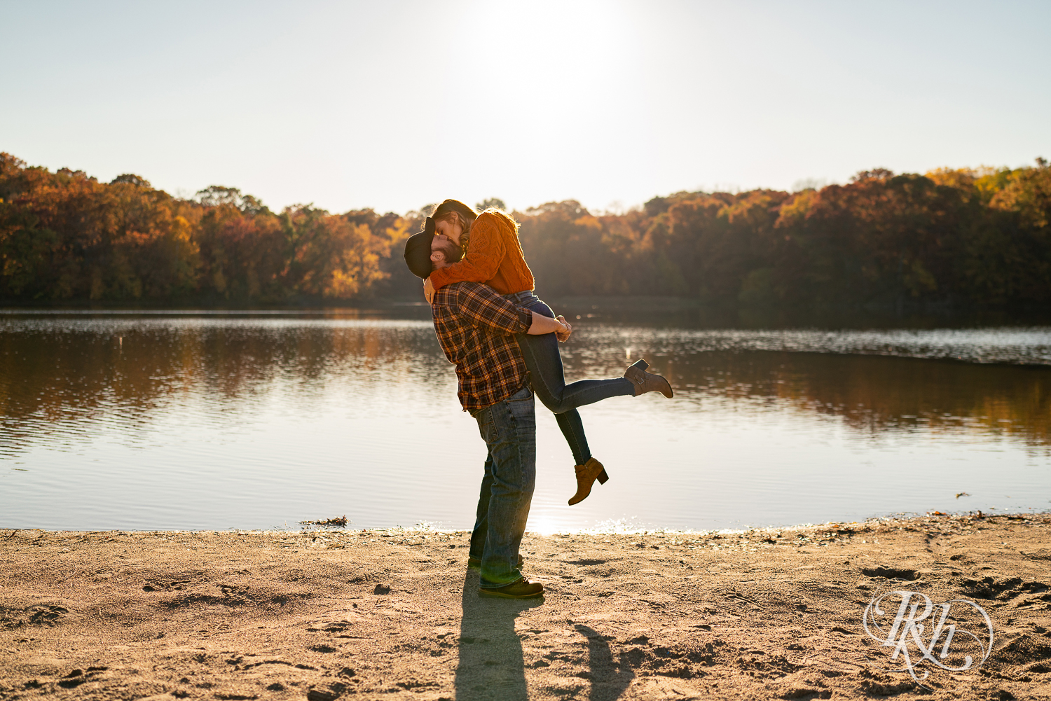 Man in flannel lifting and kissing woman in jeans and sweater on beach during sunset in Eagan, Minnesota.
