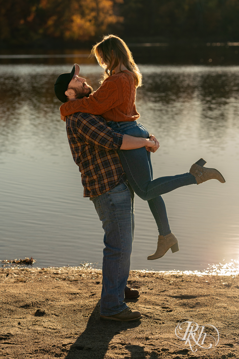 Man in flannel lifting woman in jeans and sweater on beach during sunset in Eagan, Minnesota.