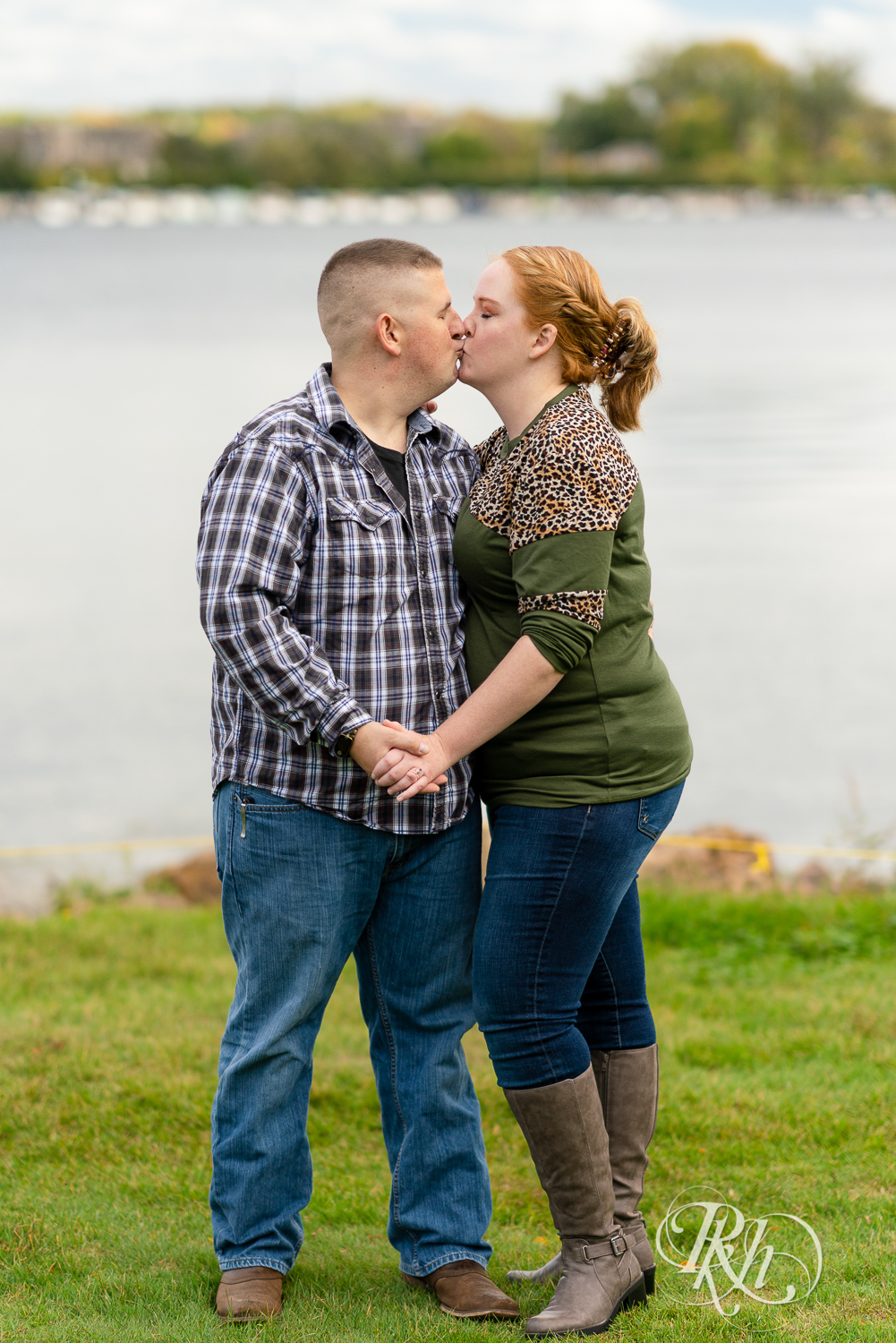 Man in flannel and jeans kisses woman in sweater and jeans in front of a lake in Excelsior, Minnesota.