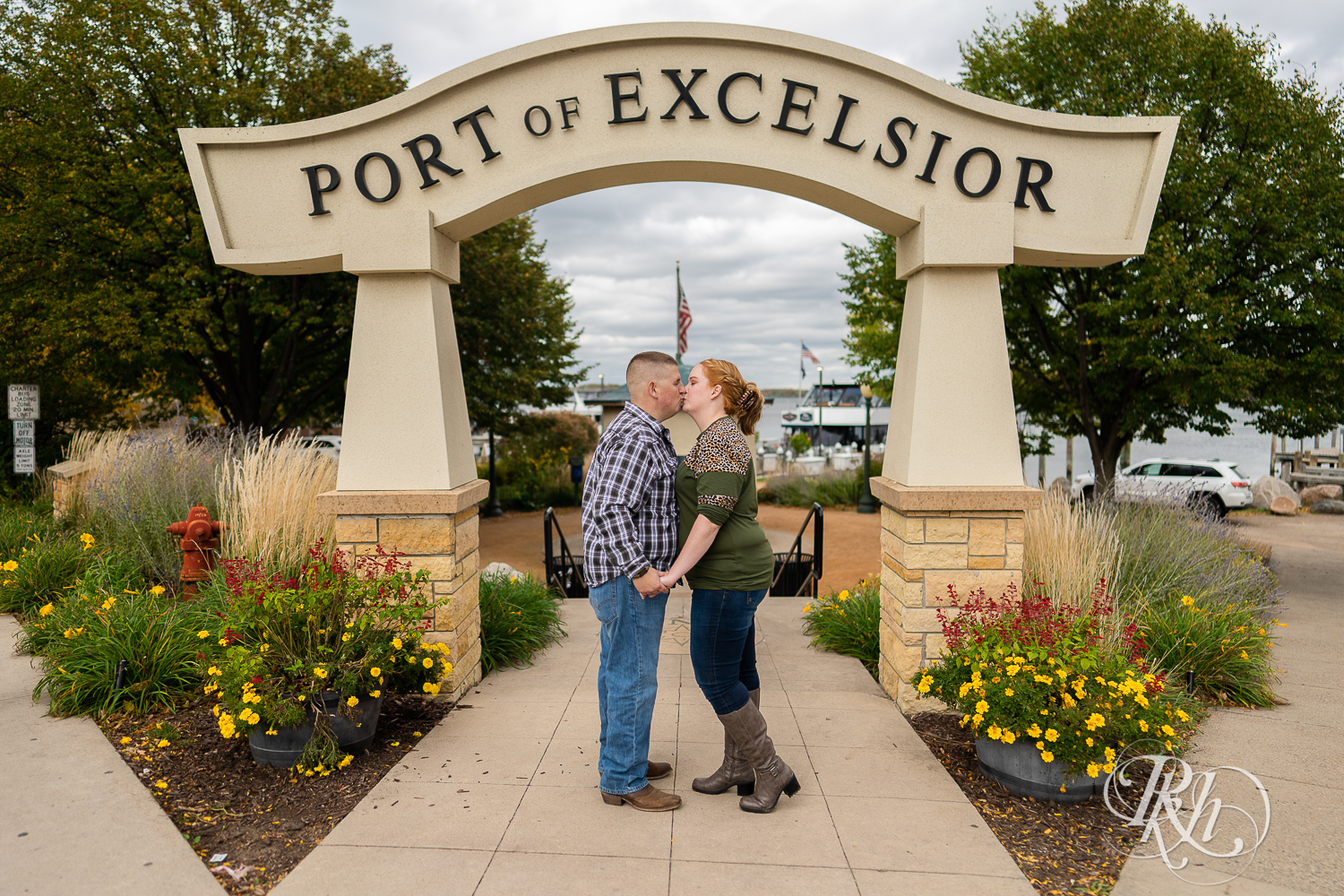 Man in flannel and jeans kisses woman in sweater and jeans in front of Port Excelsior sign in Excelsior, Minnesota.