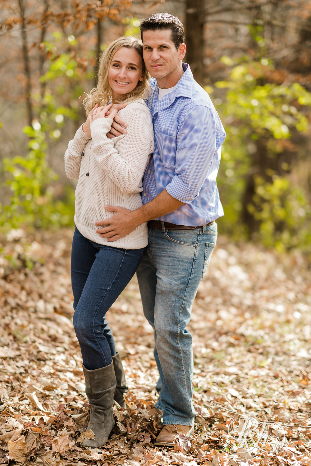 Man and blonde woman smiling in leaves during fall engagement photography session in Chaska, Minnesota.