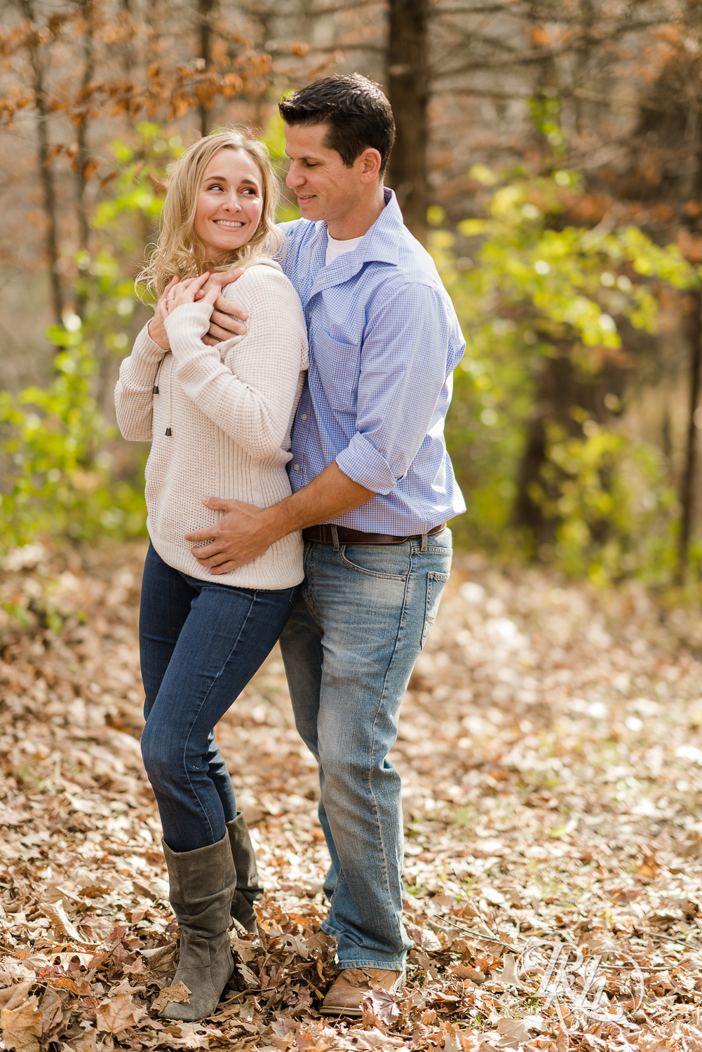 Man and blonde woman smiling in leaves during fall engagement photography session in Chaska, Minnesota.