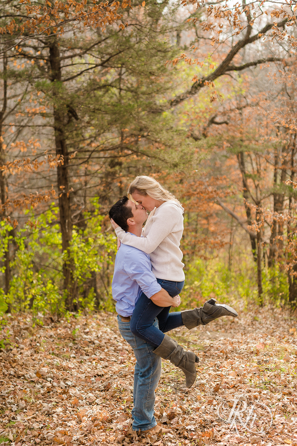 Man lifting and kissing blonde woman in leaves during fall engagement photography session in Chaska, Minnesota.