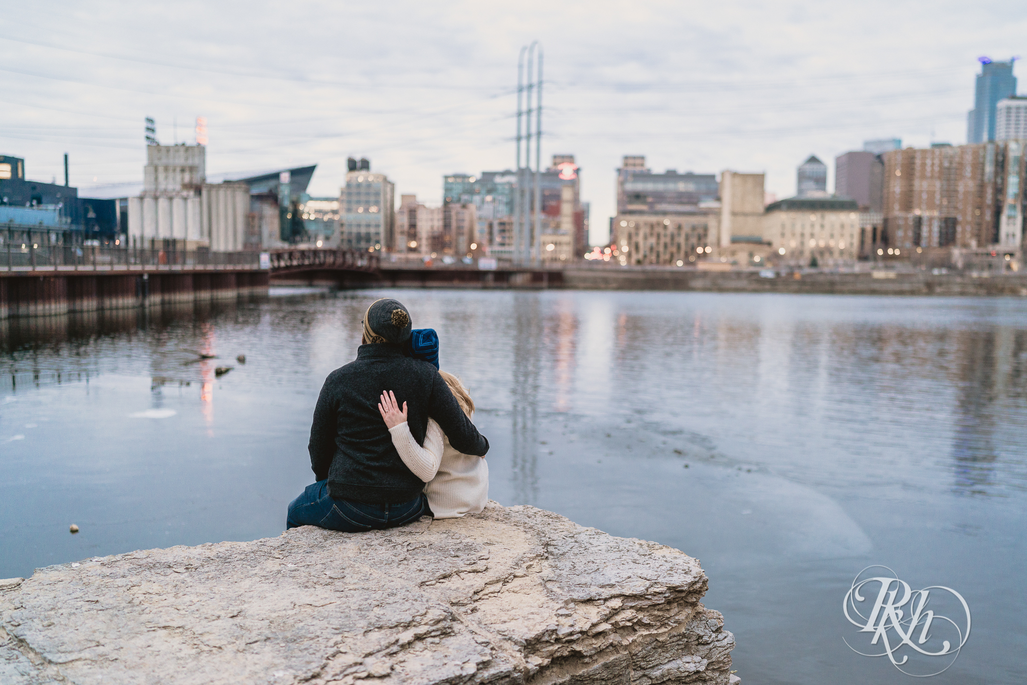 Man and woman sitting on rock facing Minneapolis, Minnesota with their arms around one another at sunrise. 