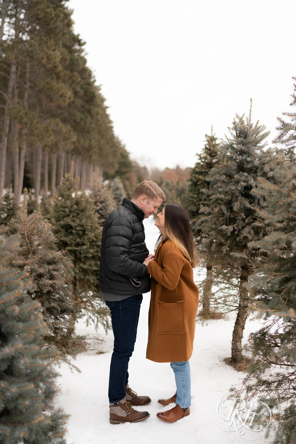 Man and woman smiling in between pine trees in the snow at Hansen Tree Farm in Anoka, Minnesota.