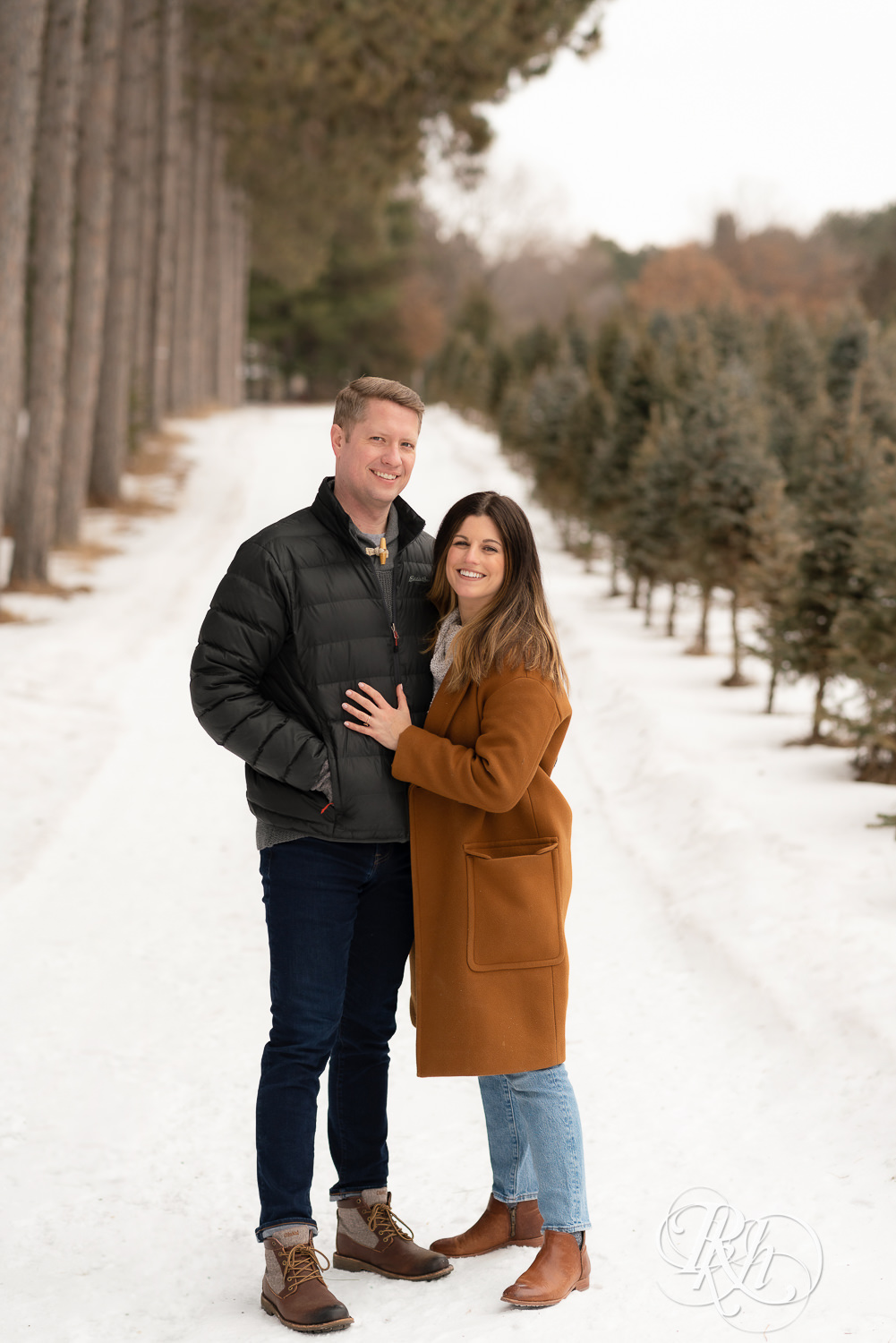 Man and woman standing in between pine trees in the snow at Hansen Tree Farm in Anoka, Minnesota.