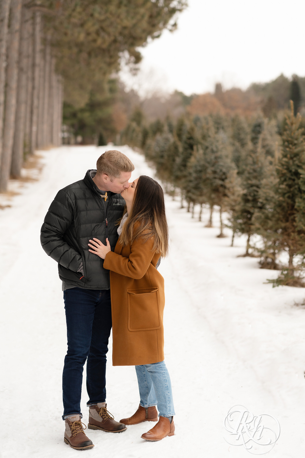 Man and woman kissing in between pine trees in the snow at Hansen Tree Farm in Anoka, Minnesota.