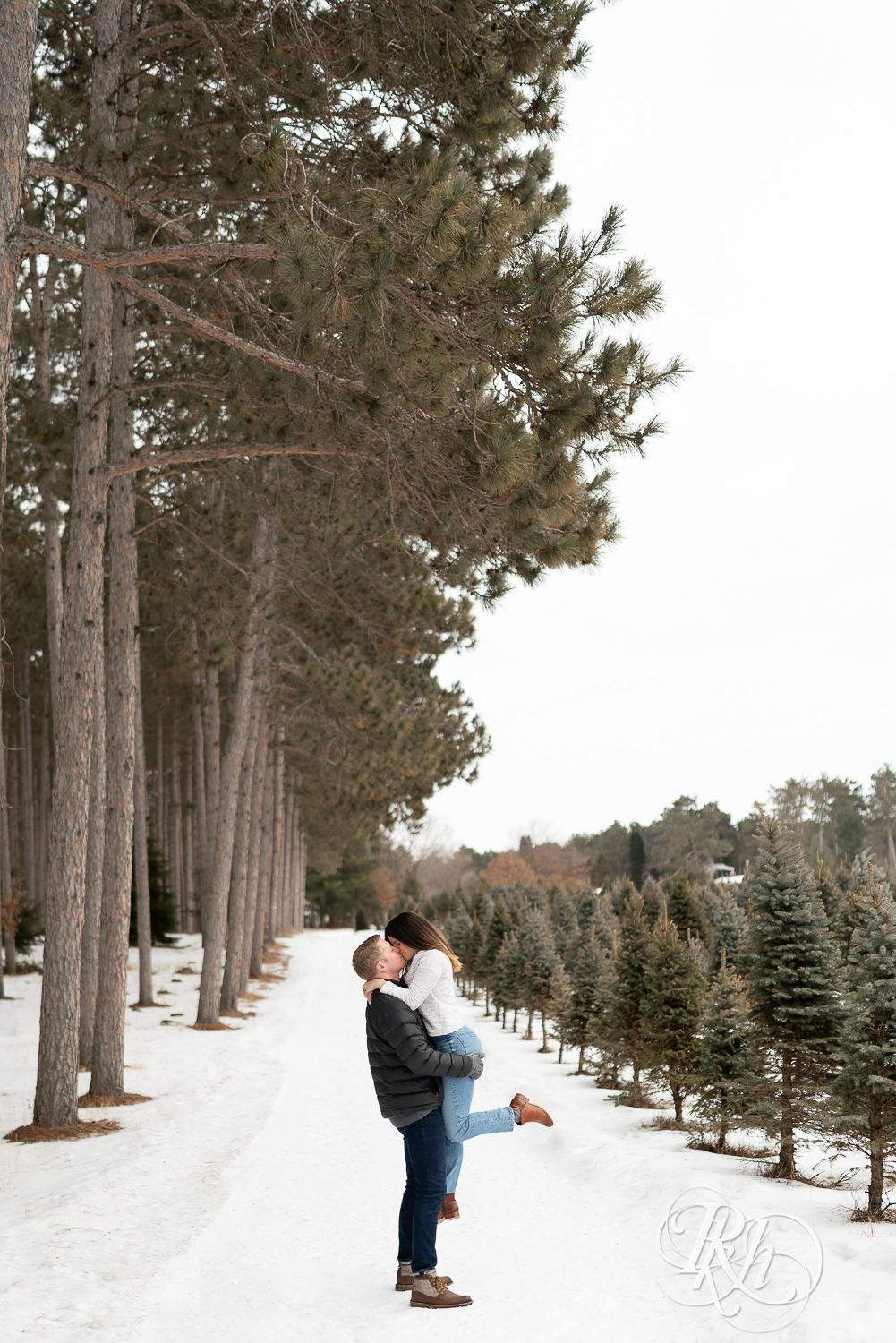 Man lifting and kissing woman in between pine trees in the snow at Hansen Tree Farm in Anoka, Minnesota.