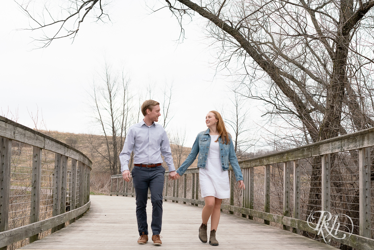Redhead couple in denim smile and walk holding hands on bridge on cloudy day in Eagan, Minnesota.