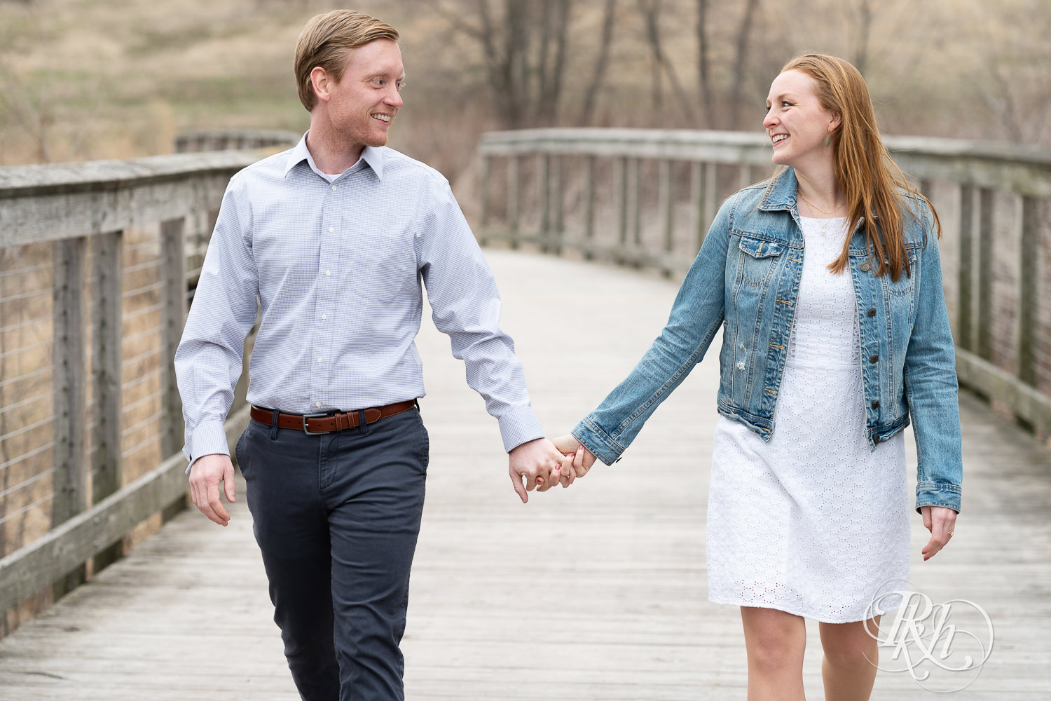 Redhead couple in denim smile and walk holding hands on bridge on cloudy day in Eagan, Minnesota.