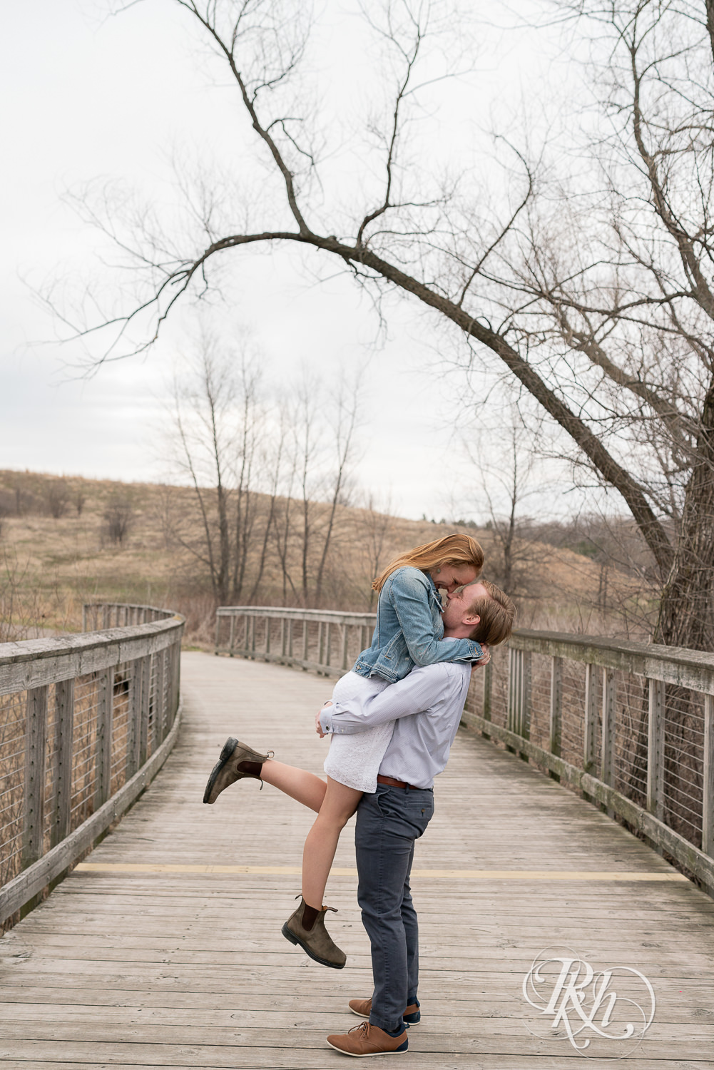 Man lifts woman in denim and dress on bridge on cloudy day in Eagan, Minnesota.