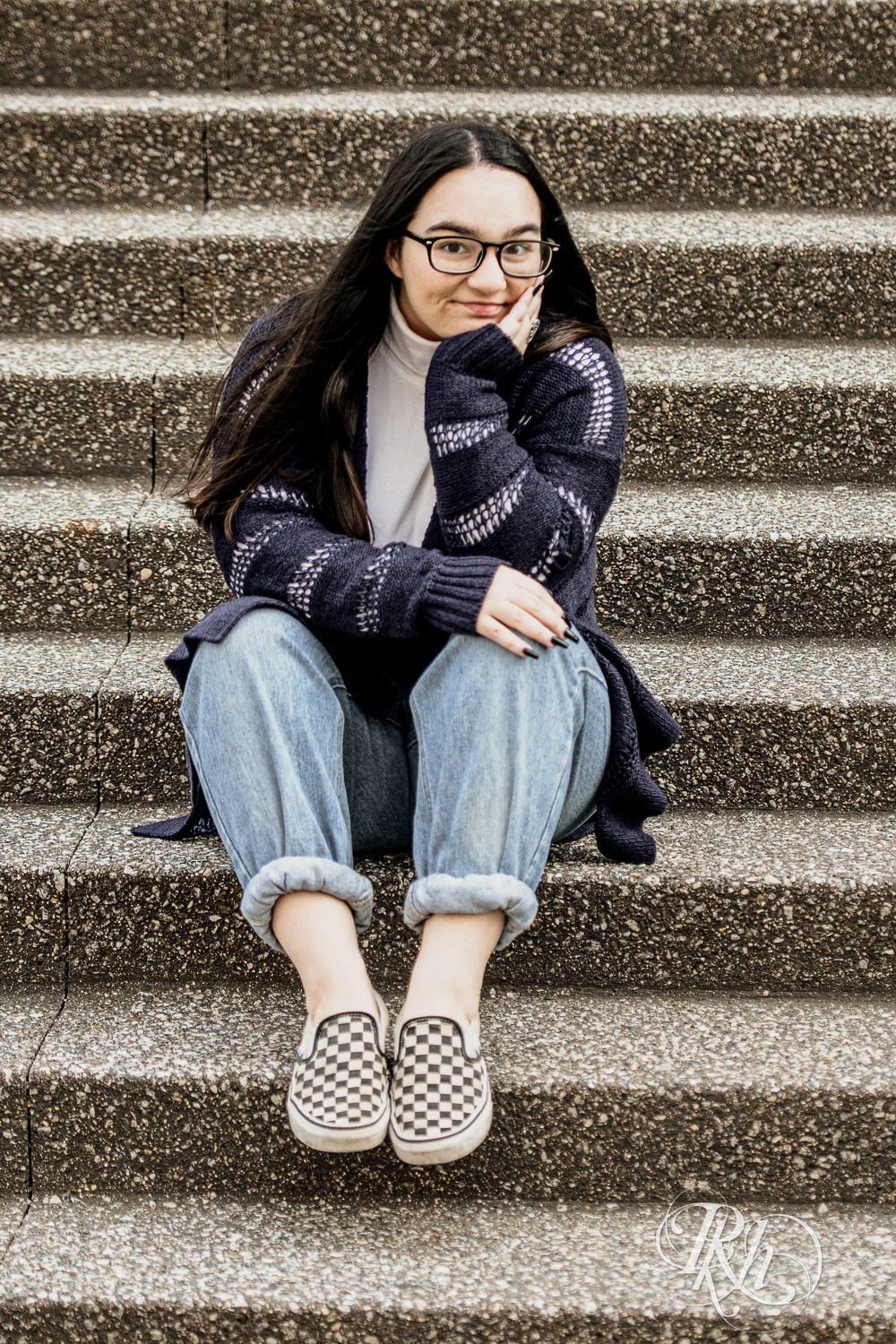 Girl dressed in glasses, sweater, and jeans seats in front of Guthrie Theater for senior photos.