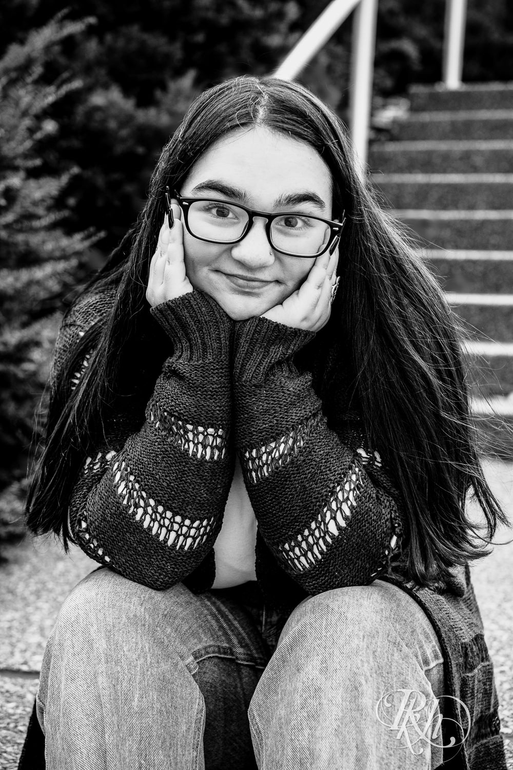 Girl dressed in glasses, sweater, and jeans seats in front of Guthrie Theater for senior photos.