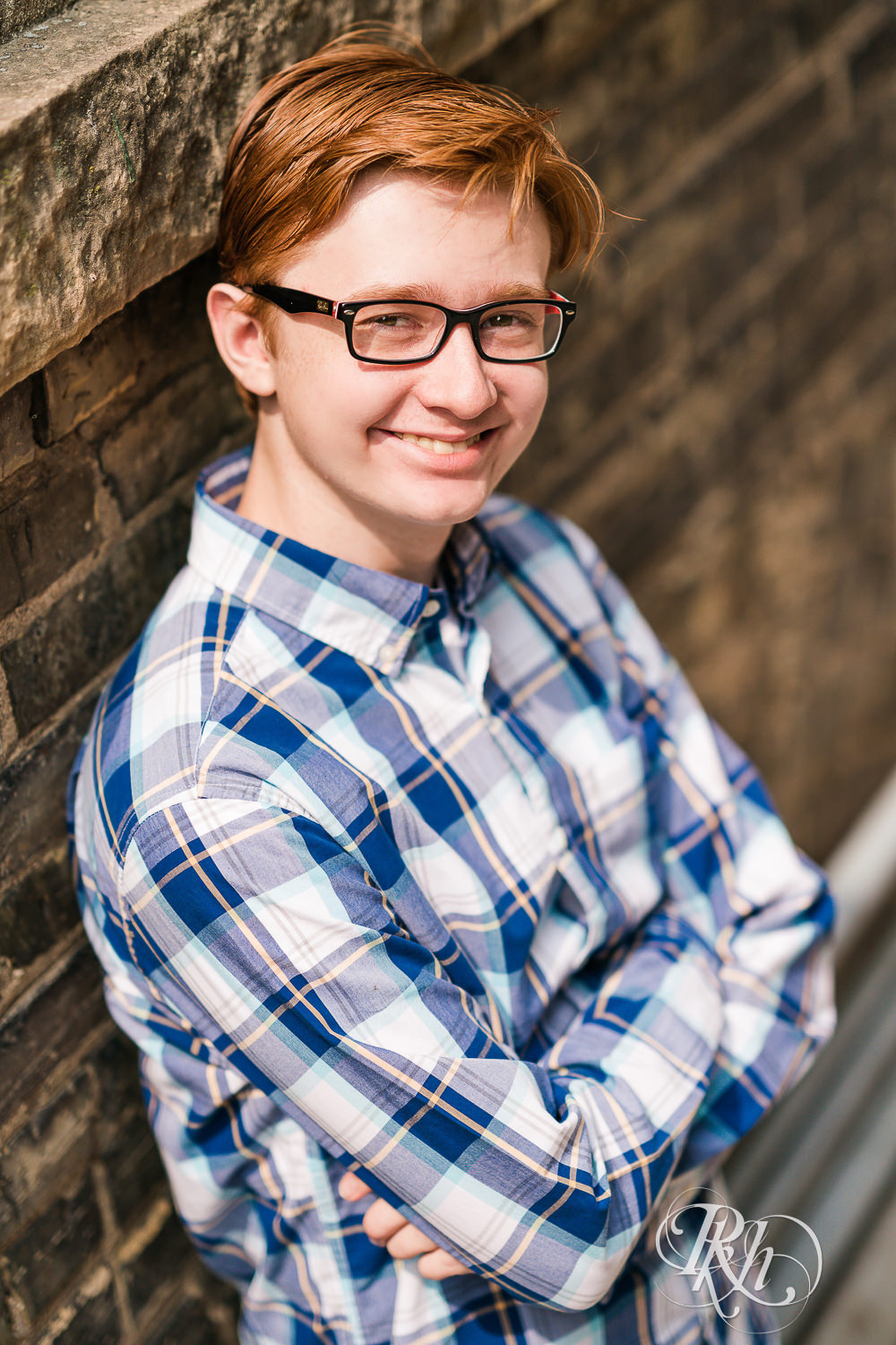 Redheaded boy in glasses and flannel leaning against brick wall for senior pictures in Minneapolis.