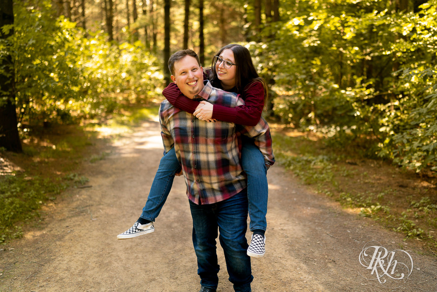Man dressed in flannel and jeans carrying woman on his back in the woods.