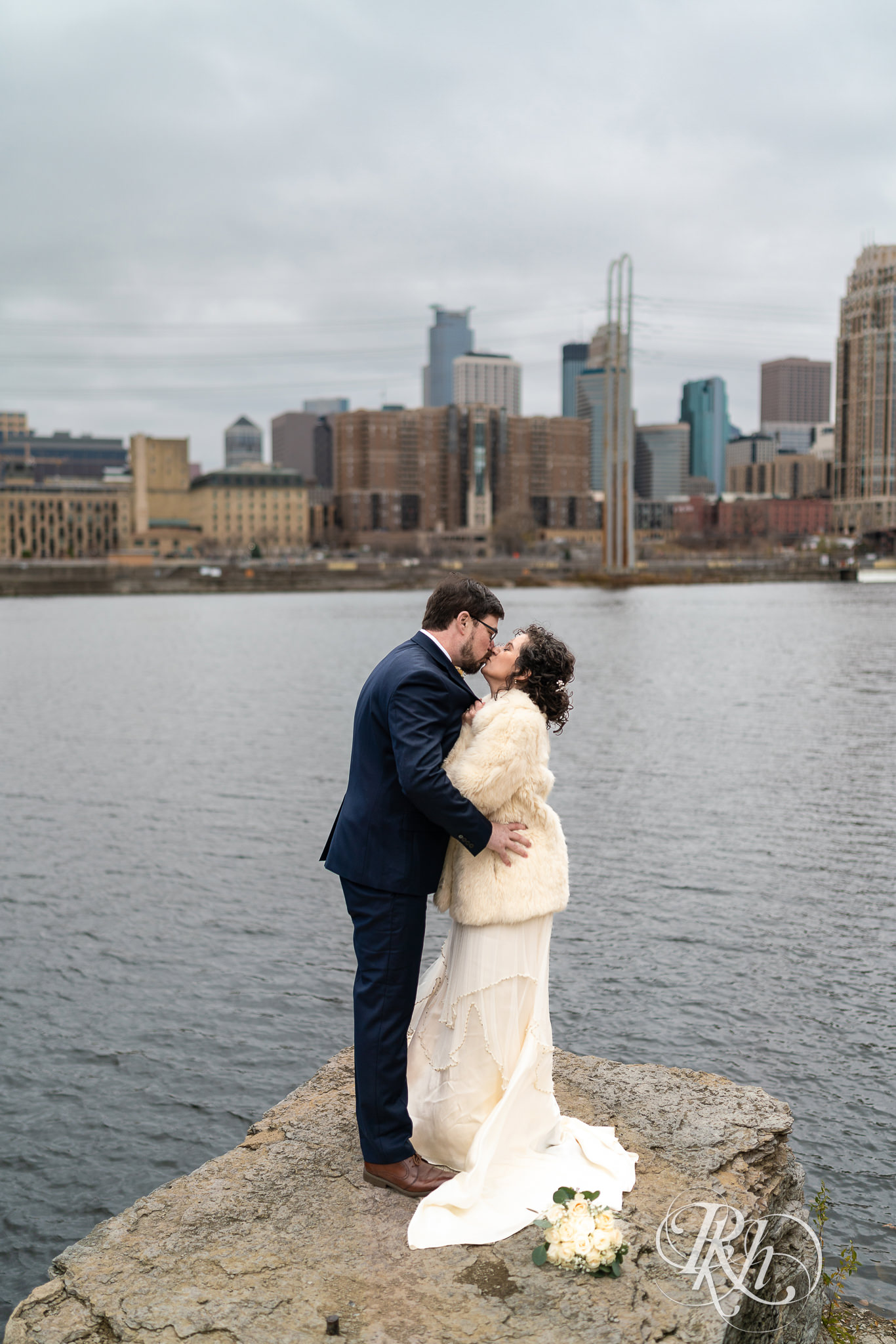 A bride and groom kiss on a rock with the Minneapolis skyline behind them. 
