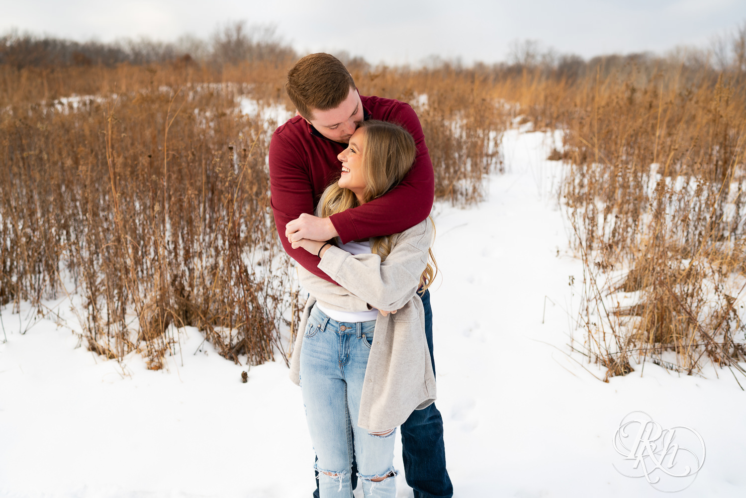 A couple stands in the snow hugging and smiling at one another. The man kisses the woman's forehead.