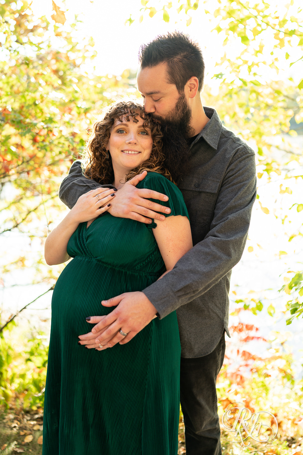 Pregnant woman in green dress and man kiss and hold belly in Lebanon Hills Regional Park in Eagan, Minnesota.
