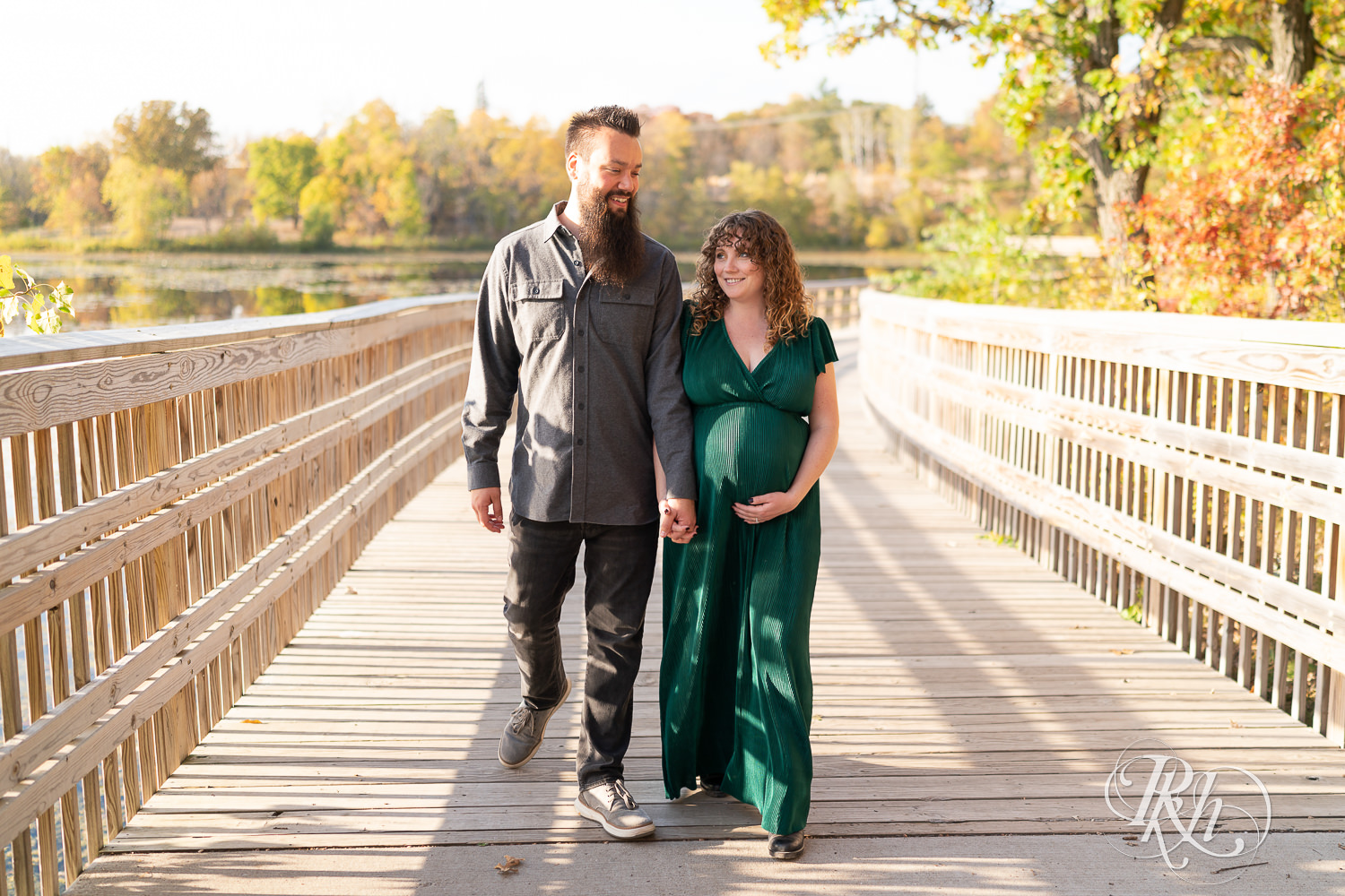 Pregnant woman in green dress and man hold belly and walk on bridge in Lebanon Hills Regional Park in Eagan, Minnesota.