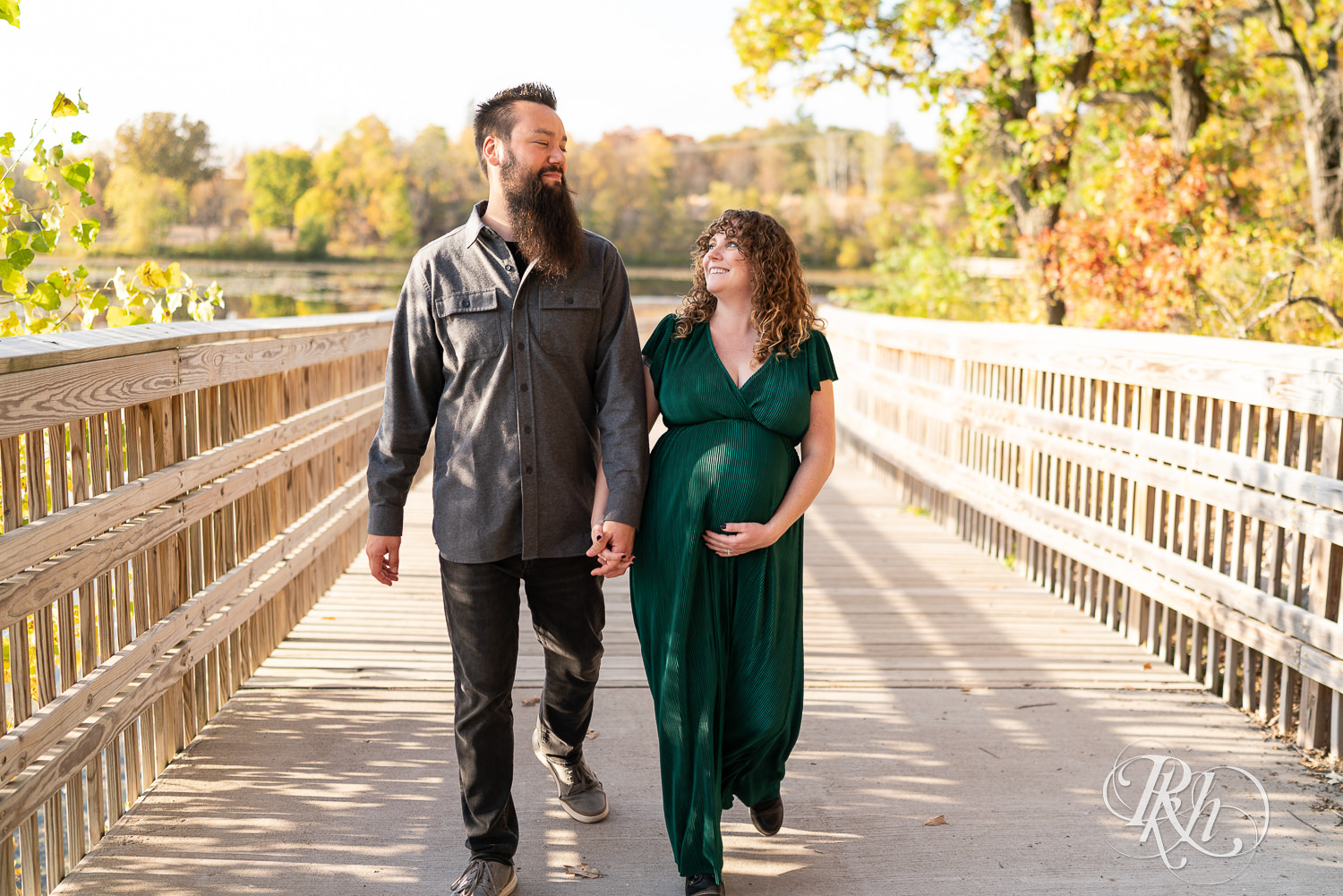 Pregnant woman in green dress and man hold belly and walk on bridge in Lebanon Hills Regional Park in Eagan, Minnesota.