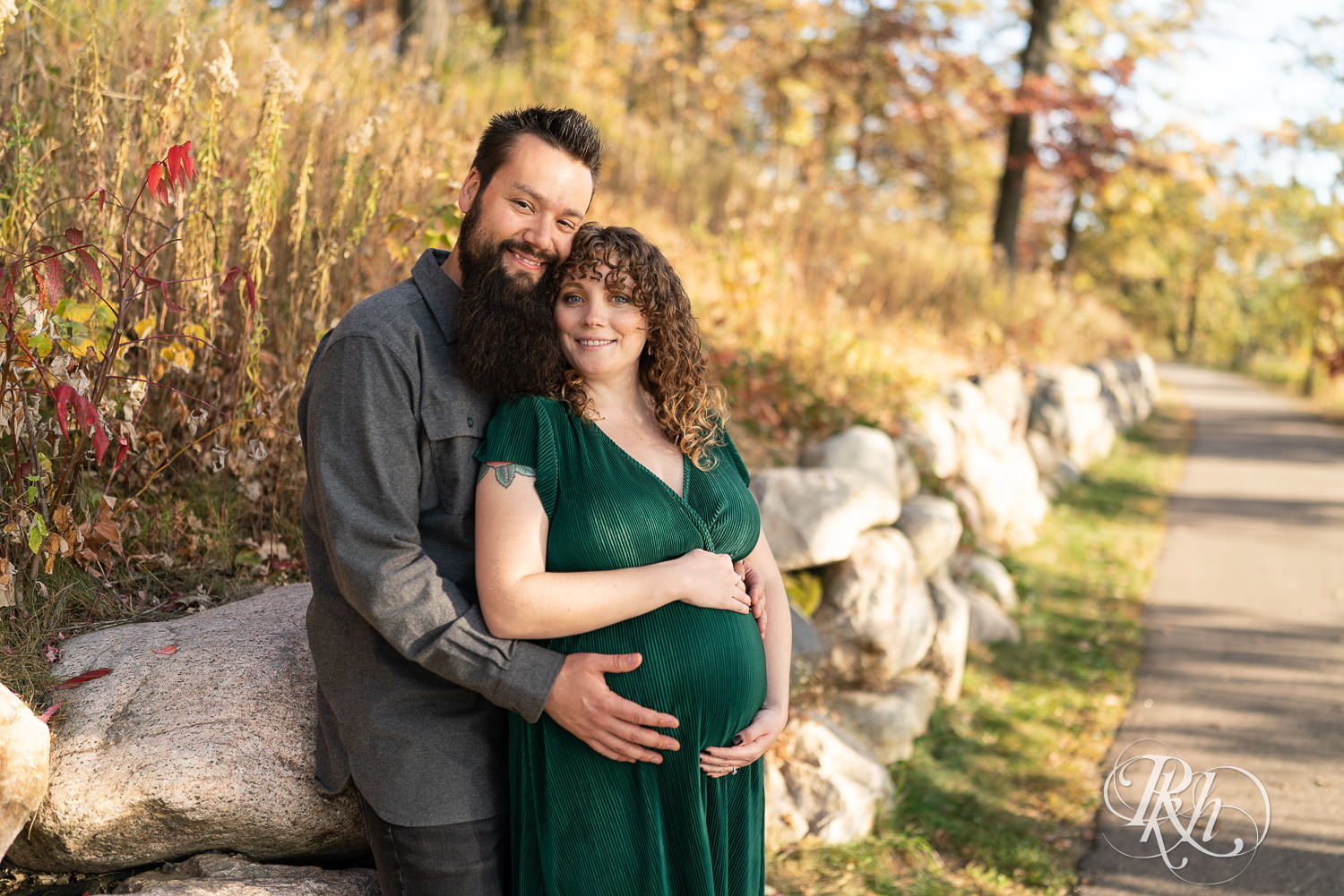 Pregnant woman in green dress and man hold belly and smile in Lebanon Hills Regional Park in Eagan, Minnesota.