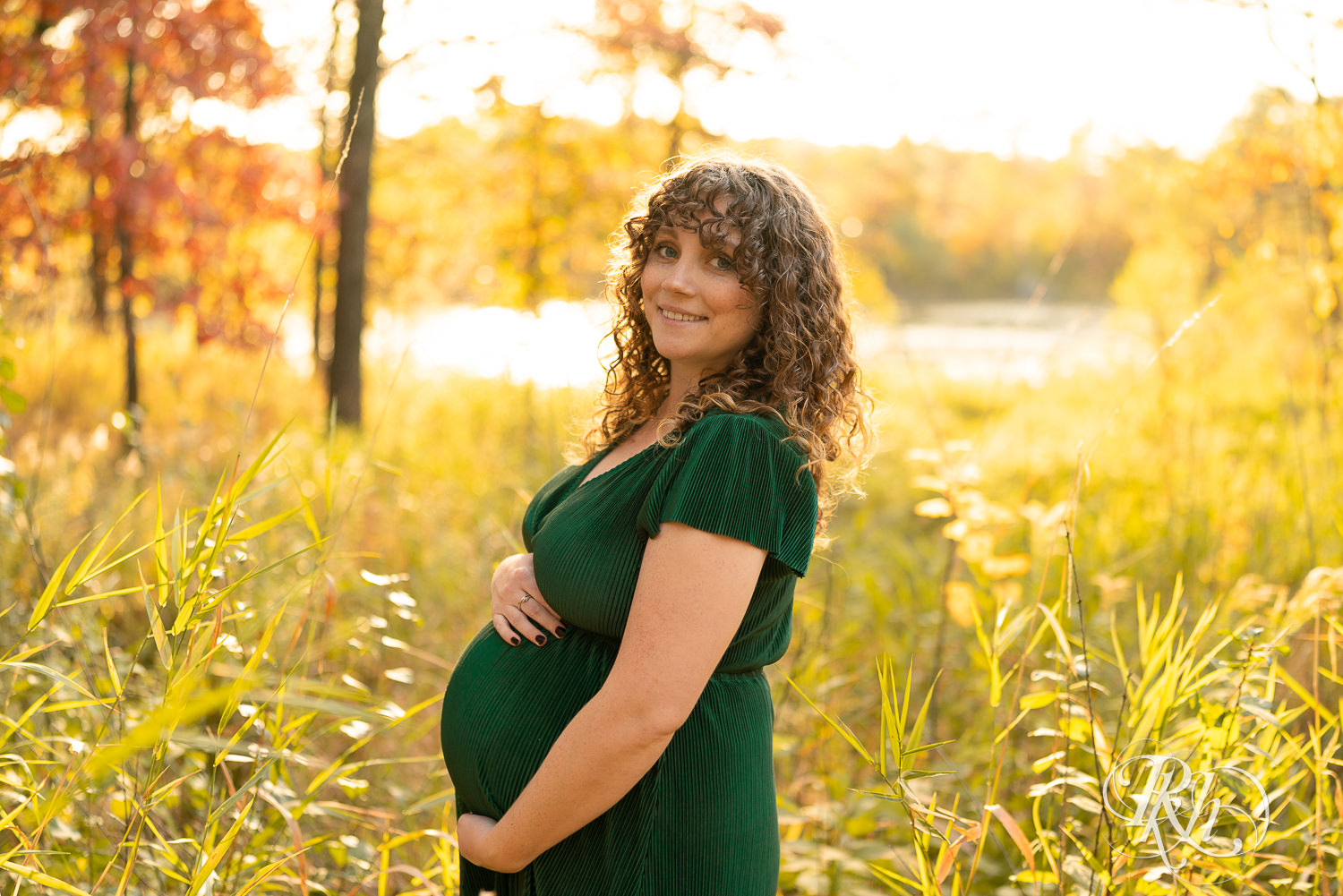 Pregnant woman in green dress holds belly in field during maternity photography in Lebanon Hills Regional Park in Eagan, Minnesota.