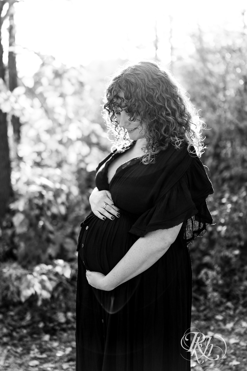 Pregnant woman in black dress in hat holds belly in field in Lebanon Hills Regional Park in Eagan, Minnesota.