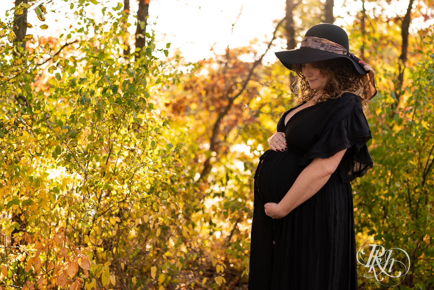 Pregnant woman in black dress and hat holds belly during sunset in Lebanon Hills Regional Park in Eagan, Minnesota.