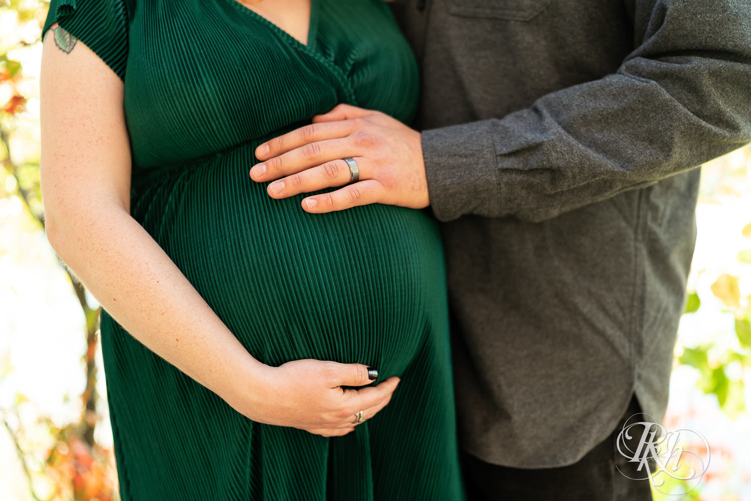 Pregnant woman in green dress and man hold belly in Lebanon Hills Regional Park in Eagan, Minnesota.