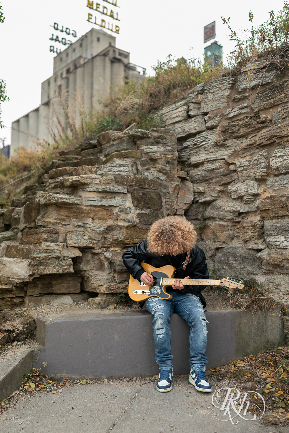Senior boy playing dressed in leather and jeans plays guitar in Mill City Ruins in Minneapolis, Minnesota.
