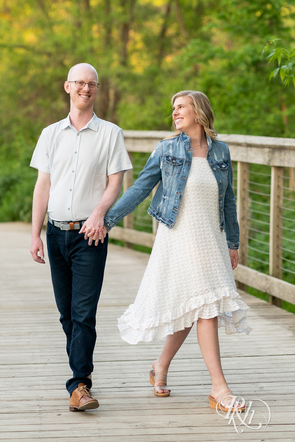 Man in white shirt and blonde woman in white dress and denim walk on bridge at sunset in Lebanon Hills Regional Park in Eagan, Minnesota.