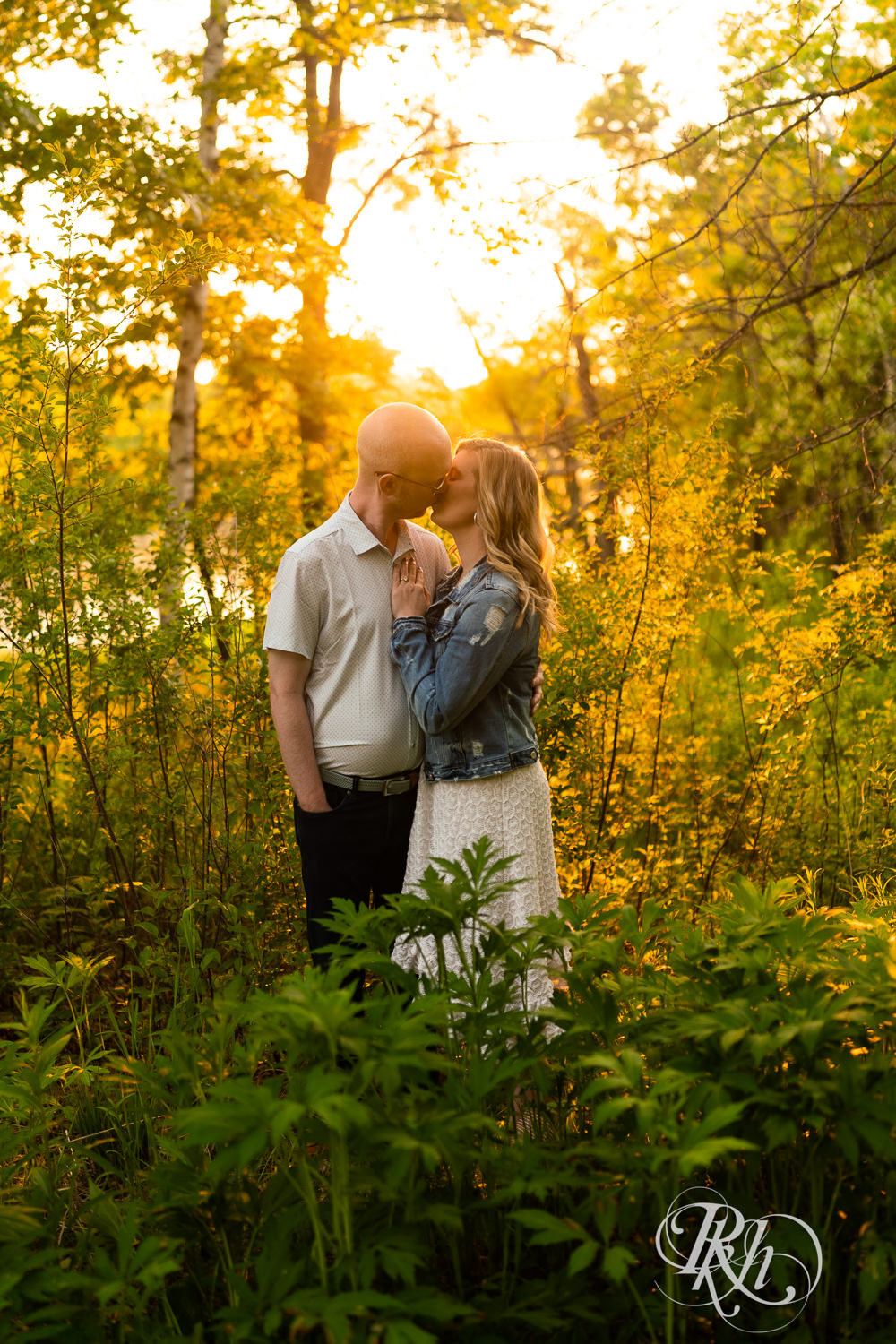 Man in white shirt and blonde woman in white dress and denim kiss at sunset in Lebanon Hills Regional Park in Eagan, Minnesota.
