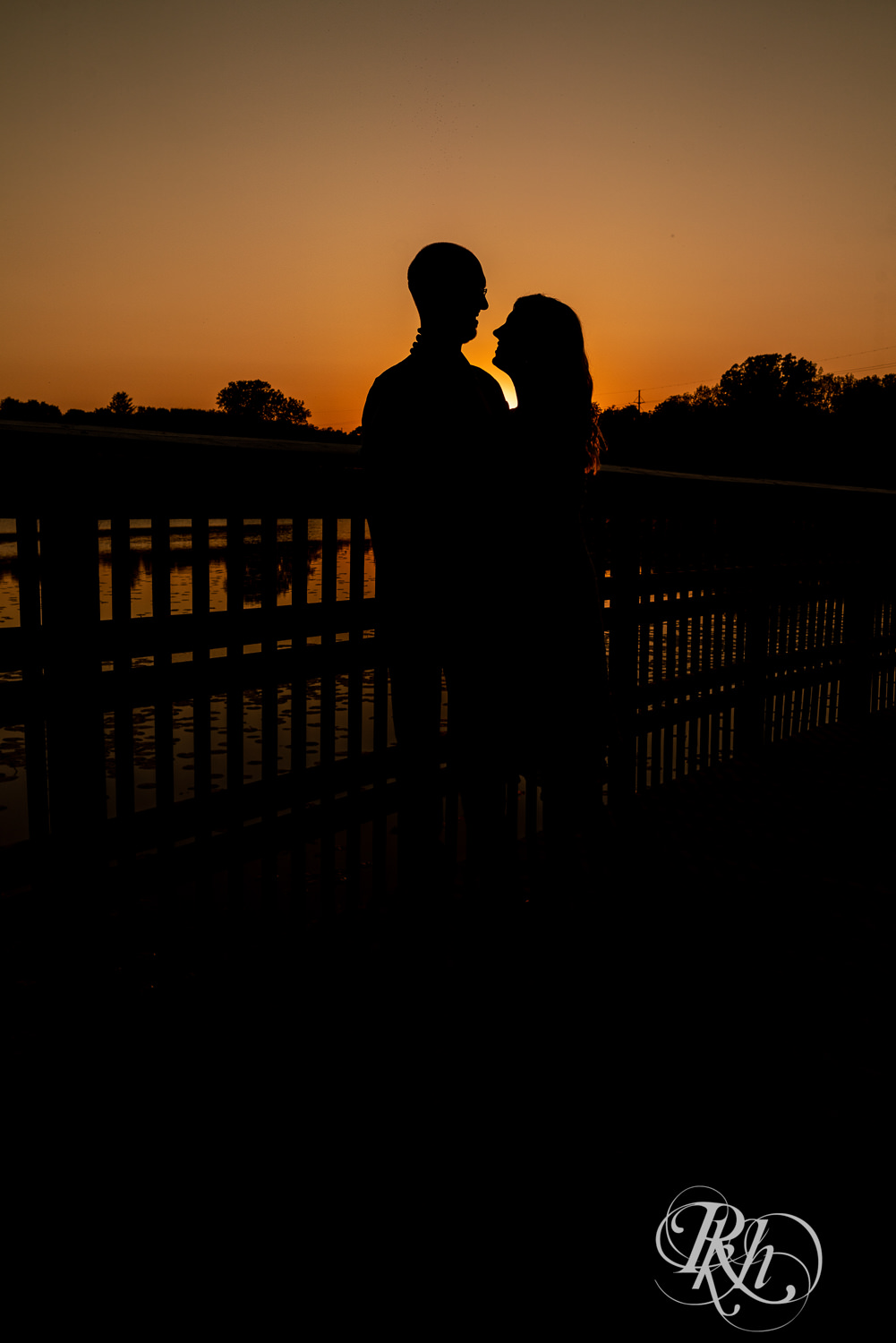 Man in white shirt and blonde woman in white dress and denim kiss in silhouette at sunset in Lebanon Hills Regional Park in Eagan, Minnesota.
