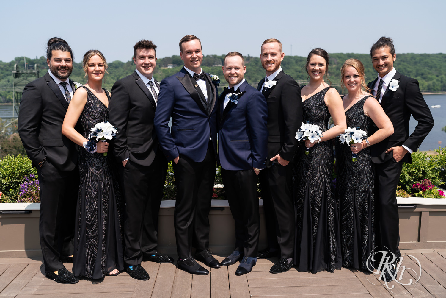 Grooms in blue tuxedos smile on rooftop with wedding party before gay wedding in Stillwater, Minnesota.
