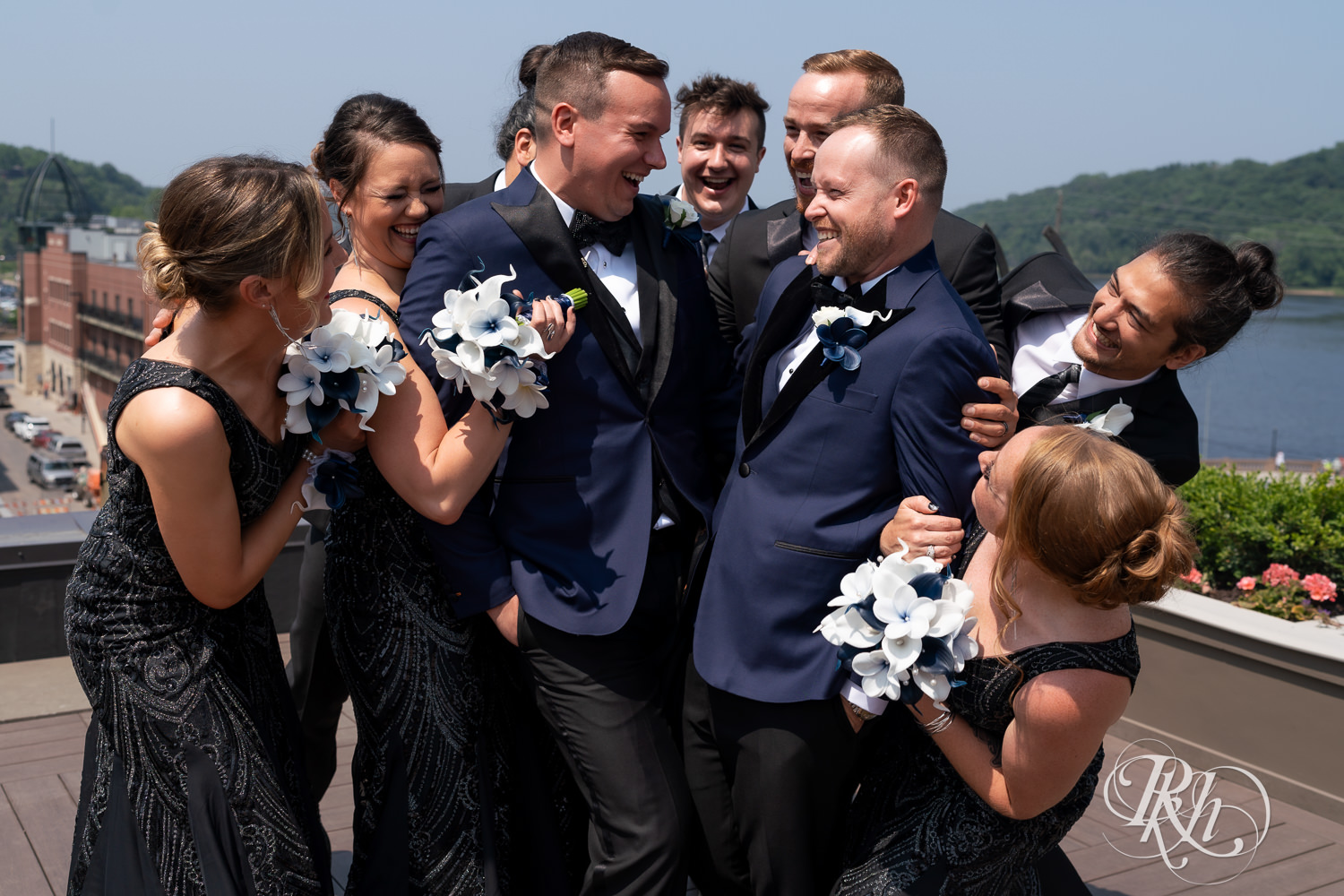 Grooms in blue tuxedos laugh on rooftop with wedding party before gay wedding in Stillwater, Minnesota.