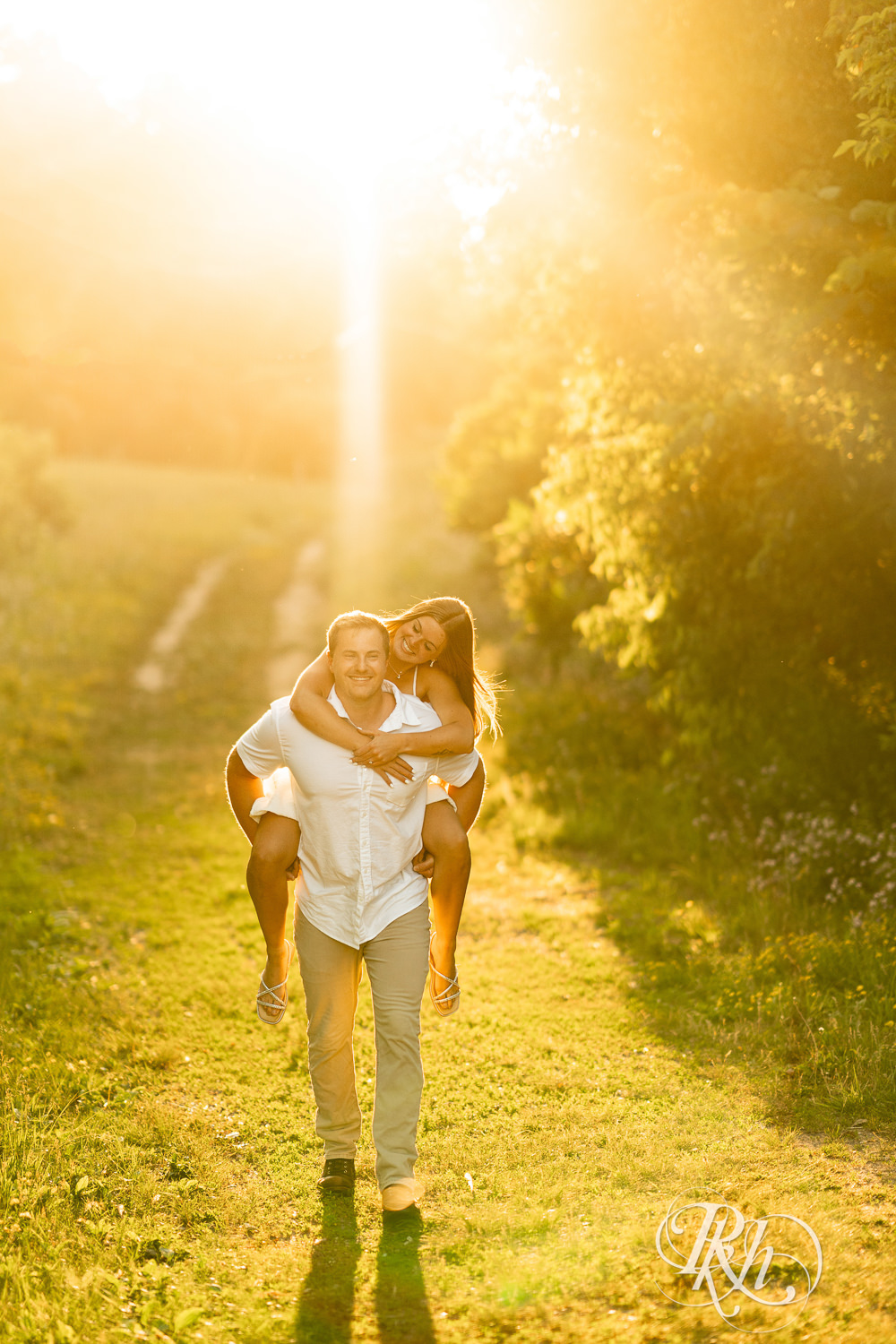 Man in white shirt carries woman in white dress on back during golden hour engagement photography at Lebanon Hills in Eagan, Minnesota.