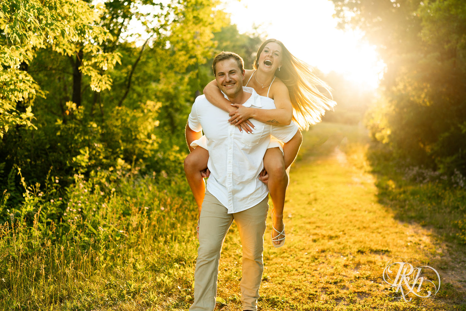 Man in white shirt carries woman in white dress on back during golden hour engagement photography at Lebanon Hills in Eagan, Minnesota.