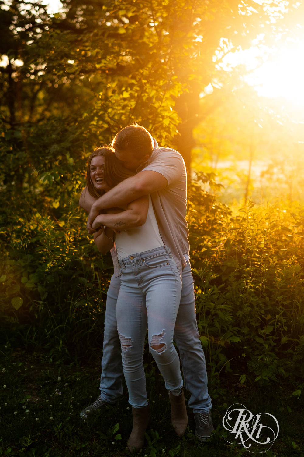 Man and woman in jeans laugh in field during golden hour engagement photography in Eagan, Minnesota.