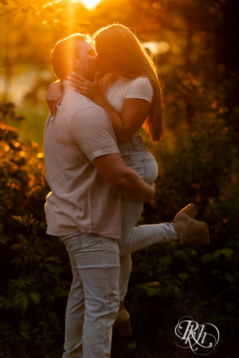 Man and woman in jeans kiss in field during golden hour engagement photography in Eagan, Minnesota.