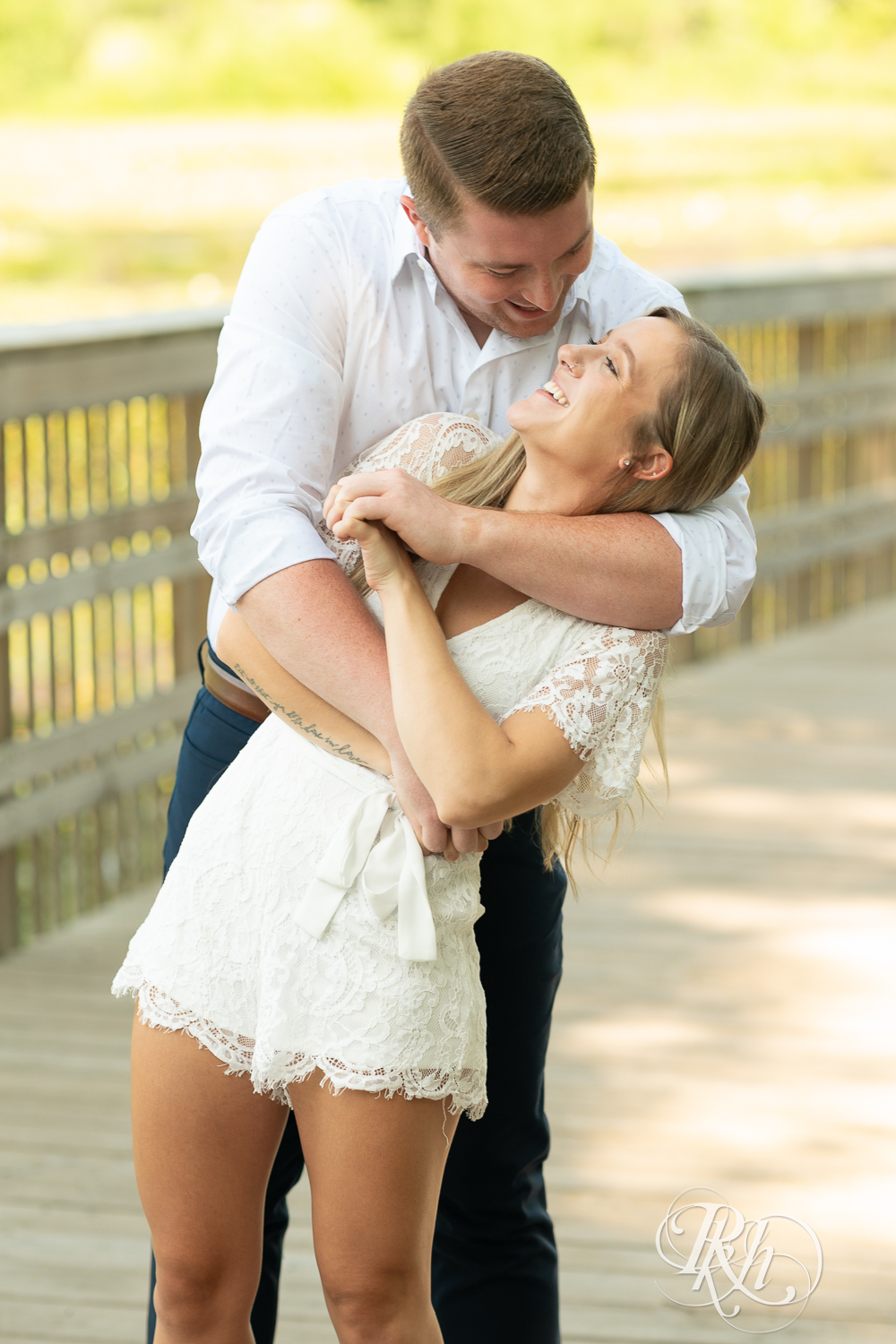 Man and woman dressed in white smile on a bridge during summer engagement photography in Minnesota.
