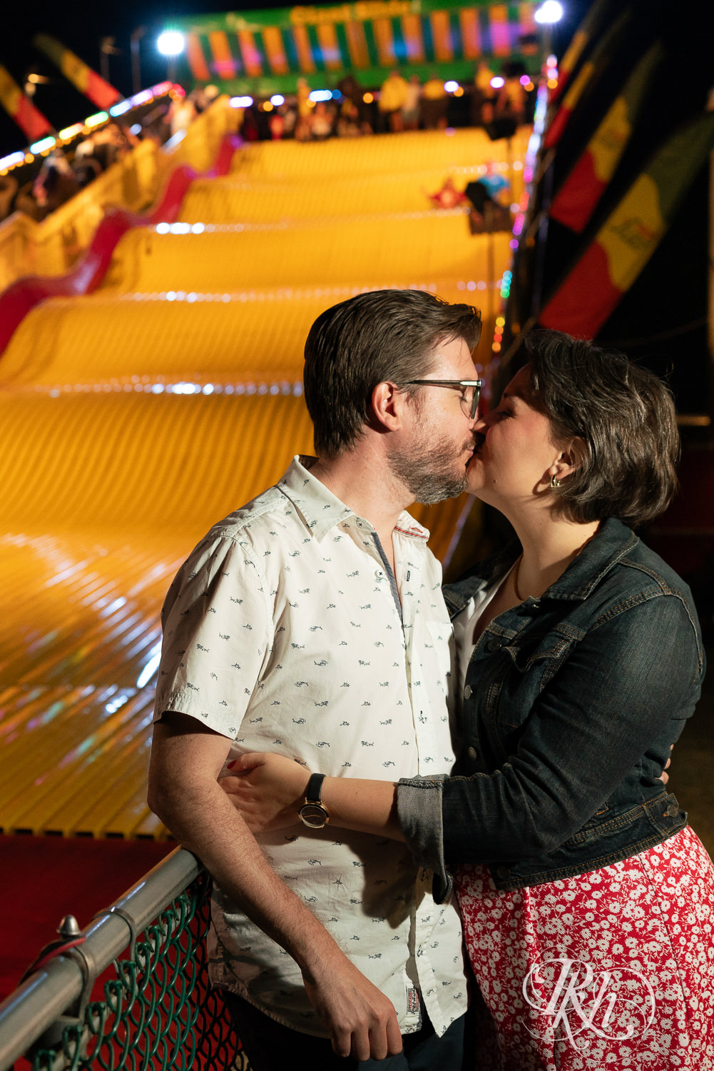Man and woman kiss in front of the giant slide during their engagement photography at the Minnesota State Fair. 