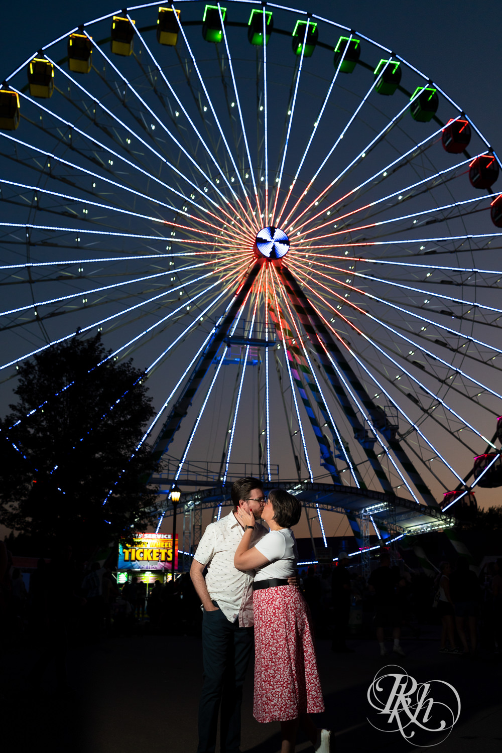 Man and woman kiss in front of the Ferris Wheel at night during their engagement photography at the Minnesota State Fair. 