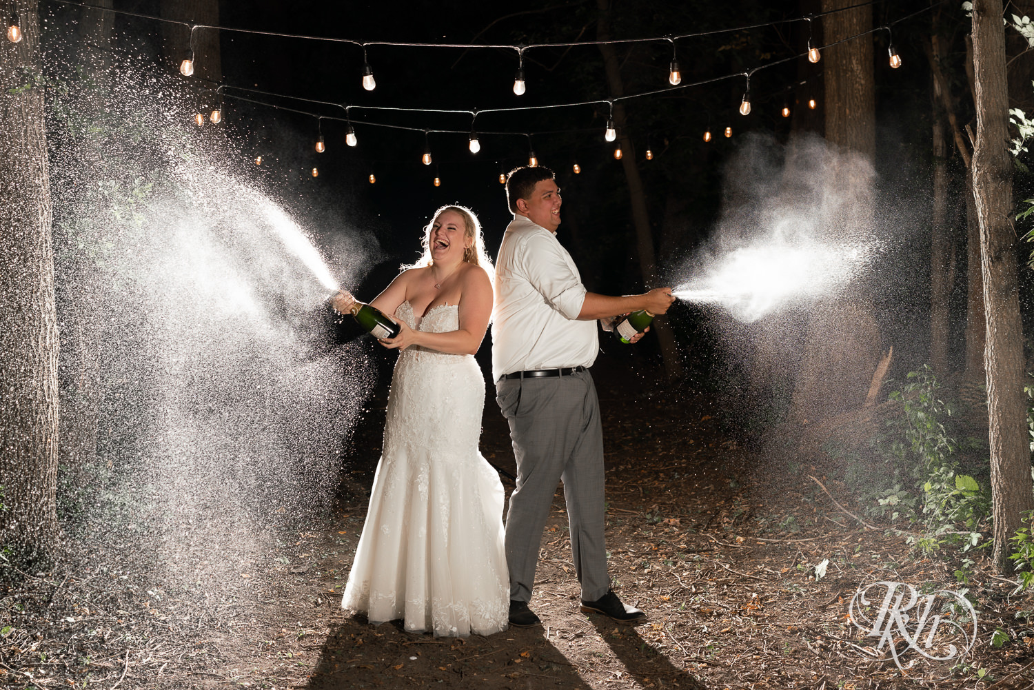 Bride and groom spray champagne while laughing under string lights in the forest.