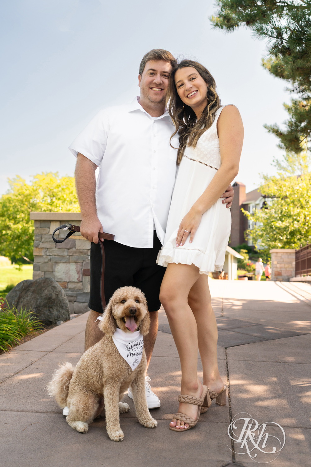 Man and woman in white dress smile with little dog during engagement photography at Centennial Lakes Park in Edina, Minnesota.