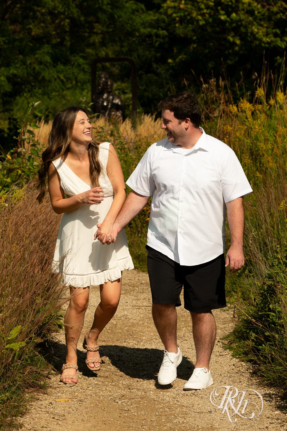 Man and woman in white dress walk together during engagement photography at Centennial Lakes Park in Edina, Minnesota.